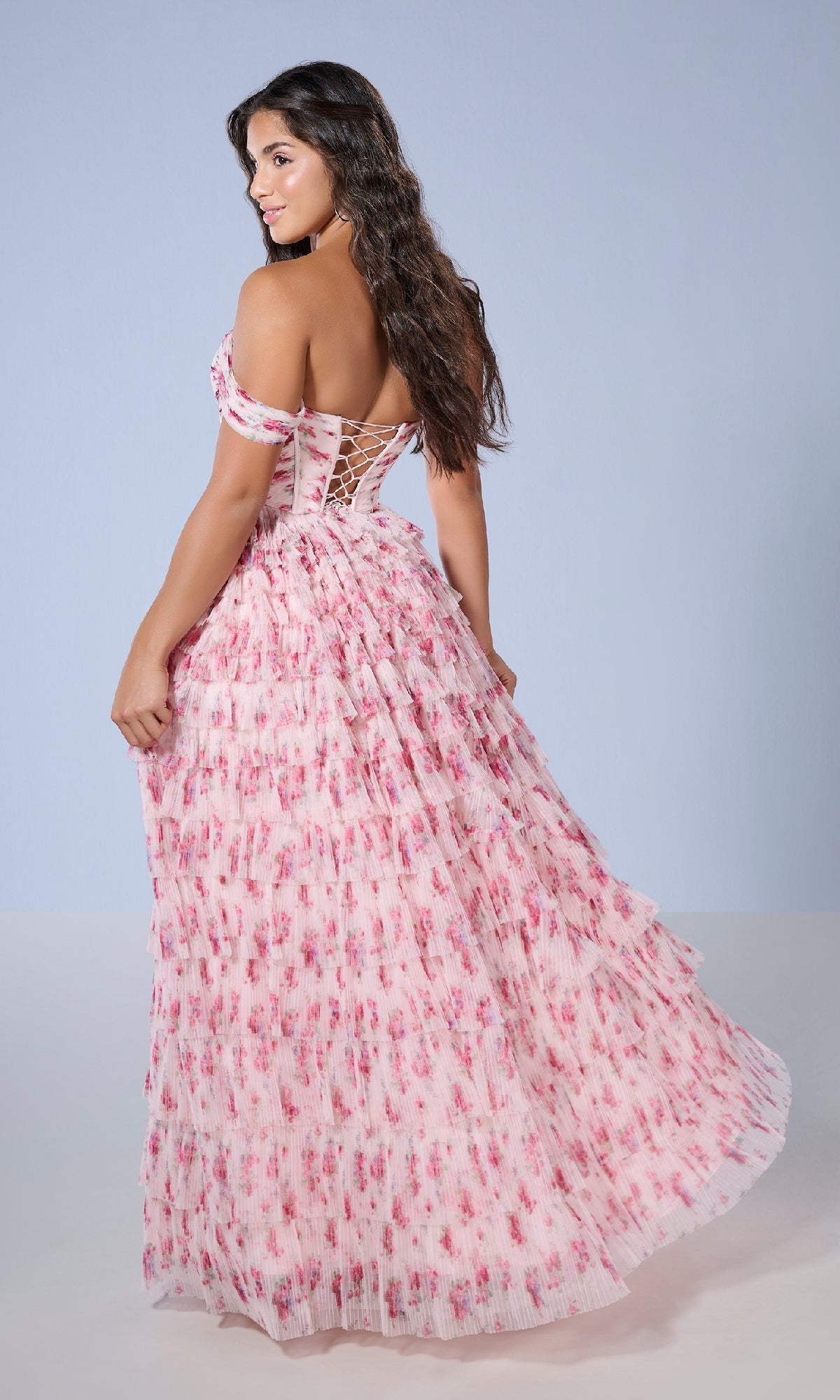 Woman wearing the Tiffany 16153 long prom dress featuring an off-the-shoulder pink floral print and corset-style lace-up back, stands looking over her shoulder against a plain light background.